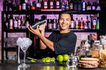 A smiling Latin American bartender mixes liquids with a cocktail shaker in front of a pub bar.