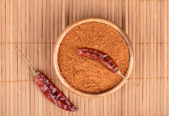 Top view of a wooden bowl with ground red pepper and hot pepper pods on a food mat.