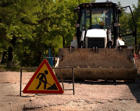 A Sign That Repairs Are Underway In The Yard, In The Background There Is An Excavator With A Bucket.