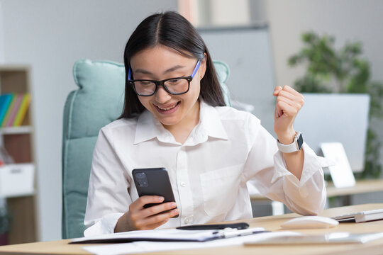 Excited beautiful young asian business woman in glasses win after achievement reading smart phone while sitting on desktop in office. She is happy, rejoices, shows a winning gesture with her hand