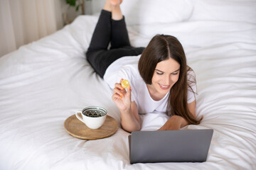 Obraz na płótnie Canvas Happy girl holding crypto coin and using laptop, lying in bed