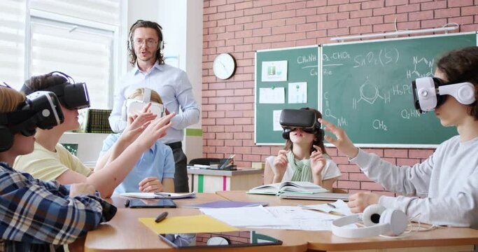 Group of young pupils in VR headsets sitting together at desk while male teacher with digital tablet in hands explaining education program. Modern technologies during interactive lesson at school.