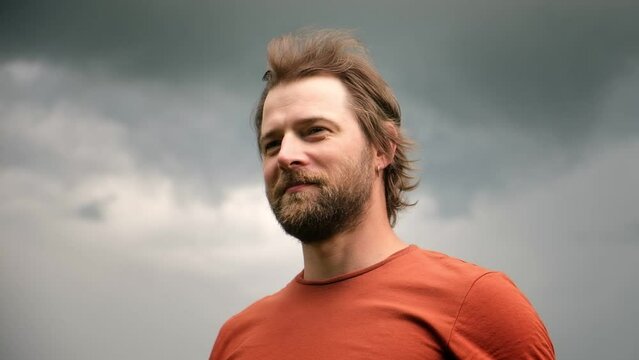 Portrait of handsome young man in orange t-shirt outdoors. Male with long hair and beard feel happy, calm and serious. Dramatic dark sky background. Man looking away into distance. Diversity People.
