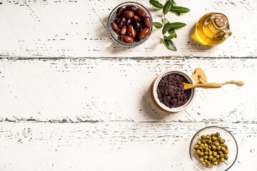Olive pate in ramequin bowl and ingredients - olive oil, olives, capers on white wooden background with olive branch