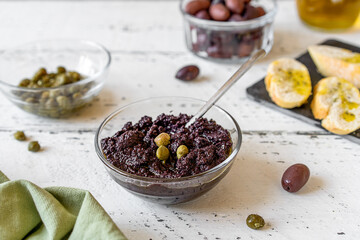 Olive pate in bowl and basic ingredients - olive oil, olives and capers on white wooden background