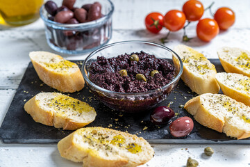 Olive pate in bowl with crostini on black stone cutting board and basic ingredients aside - olives,...