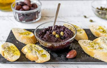 Olive pate in bowl with crostini on black stone cutting board and basic ingredients aside - olives, oil, capers. - obrazy, fototapety, plakaty