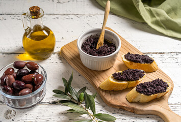 Olive pate in bowl and spread on bread slices on wooden cutting board. 