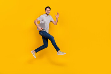 Fototapeta na wymiar Full body profile photo of cool brunet millennial guy run with laptop wear t-shirt jeans footwear isolated on yellow background