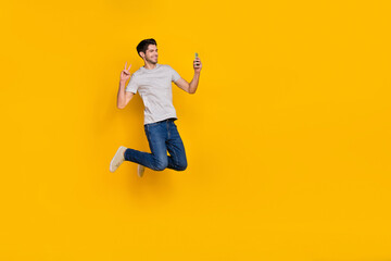 Fototapeta na wymiar Full body photo of cute brunet young guy jump do selfie wear t-shirt jeans footwear isolated on yellow background