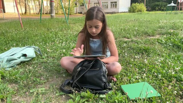 A little girl is playing a tablet while sitting alone in the street. the girl is doing lessons in nature, it's time to go to school, the girl maintains her blog, communicates with subscribers or frien