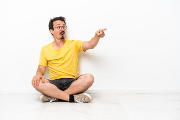 Fototapeta premium Young caucasian man sitting on the floor isolated on white background pointing away