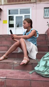 A little girl is playing a tablet while sitting alone in the street. the girl is doing lessons in nature, it's time to go to school, the girl maintains her blog, communicates with subscribers or frien