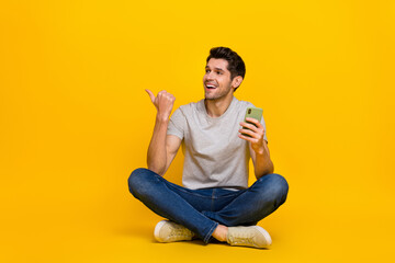 Full size photo of impressed brunet young guy sit hold telephone look promo wear t-shirt jeans shoes isolated on yellow background