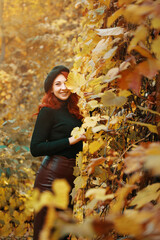 Cheerful red-haired girl covers eye with autumn leaf. Beautiful woman in fall park on walk. Female with black beret, in knitted sweater and leather skirt. Romantic model smiles and looking at camera.