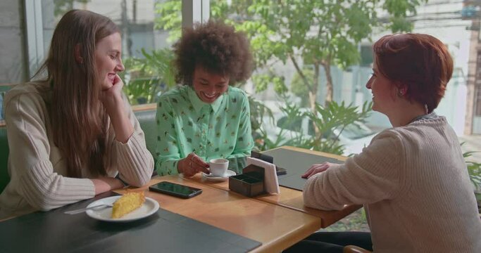 Happy diverse female friends hanging out together at coffee shop. Three young women talking inside cafe smiling and laughing