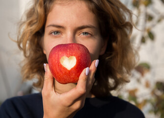 young woman holds in front of face, showing only her eyes, large red apple with heart carved on it....