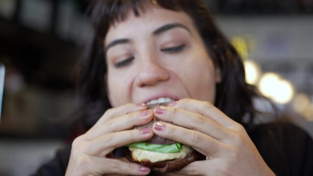 Closeup hand holding hamburguer. Young woman eating burger at restaurant. Girl takes a bite of fast food