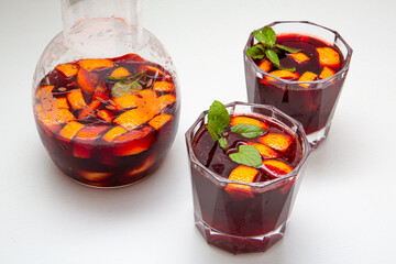 Full jar and two glasses of homemade sangria cocktails from red wine, ice and mint inside with slices of orange and lemon on a white table. 
