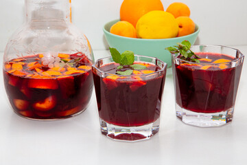 Full jar and two glasses of homemade red wine sangria cocktails ice and mint inside, with a bowl of orange, lemon, apricot background on a white table. copyspace