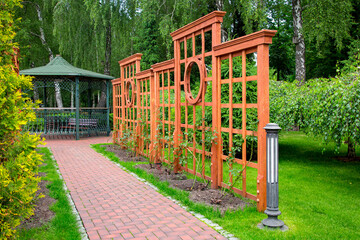 Fototapeta na wymiar Vertical wooden pergola with square pattern in growth roses garden with stone brick way and ground lantern in backyard with different trees and lawn with iron forged gazebo on background, nobody.