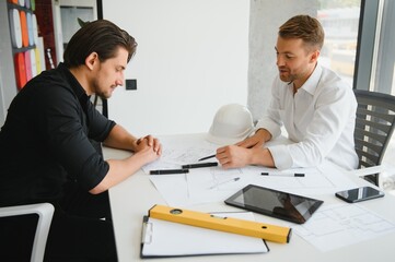 Two young architects with blueprints of a house standing in office, talking.