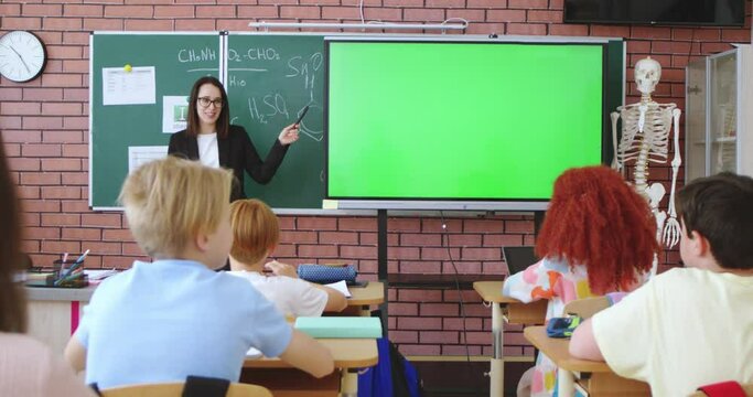 Caucasian female teacher using interactive digital board with chroma key display while explaining education program to pupils during lesson. Elementary school class with modern education.