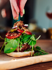 Female hand dressing a yummy burger with meat, salad and other ingredients on a wooden surface