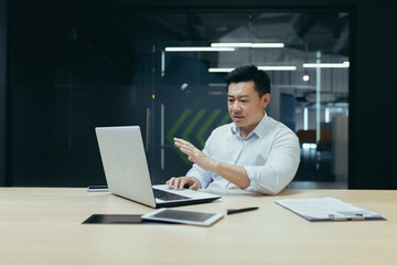 A serious young man, an Asian businessman, a freelancer, talks via video call from a laptop with clients and partners. Sitting at a desk in a modern office