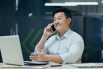 Online store. A young Asian salesman talks on the phone with customers, offers them to buy goods. Sitting behind a desk in a modern office.
