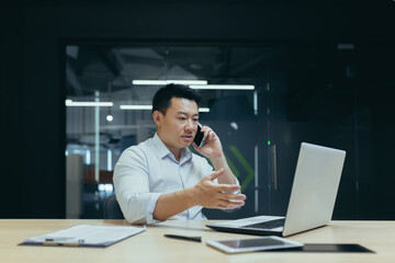 Worried and serious young male Asian businessman talking on the phone in the office. Sitting at a table with a laptop and documents. Trying to solve problems