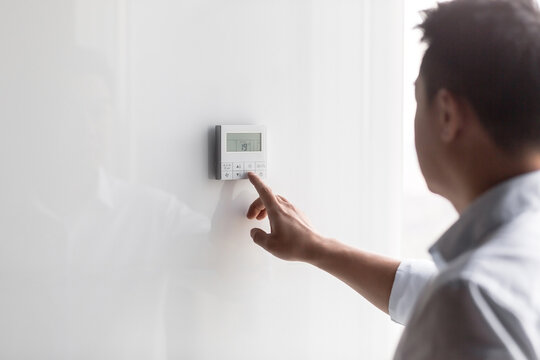 Close-up photo. The hand of a young man in a white shirt turns on the control buttons of the air conditioner hanging on the wall. Standing on the right