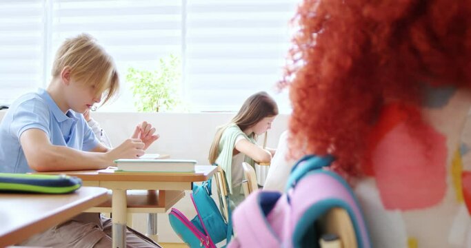 Focused caucasian children sitting at desks and writing control work. Side view of smart female teacher in formal wear controlling process of children's examination at school.