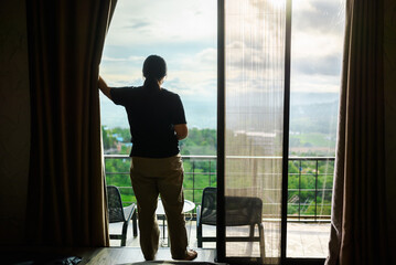 A woman open the curtain of the hotel room at the balcony to let the light in and see the mountain view