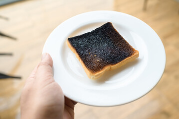 Hand holding the white dish with burnt toast on it, over heat, and mistake in the morning, danger of charred food