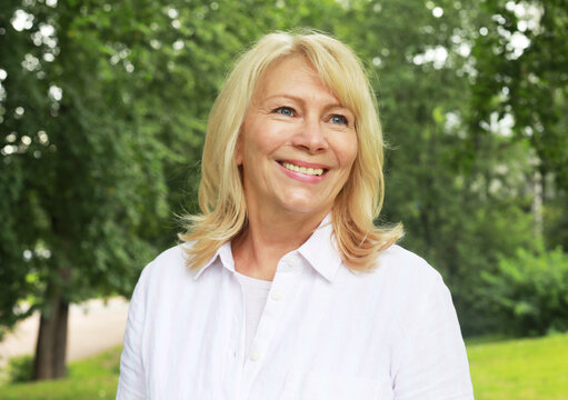 Beautiful blonde woman of sixty years old in a summer park, close-up portrait. The lady smiles and feels happy.