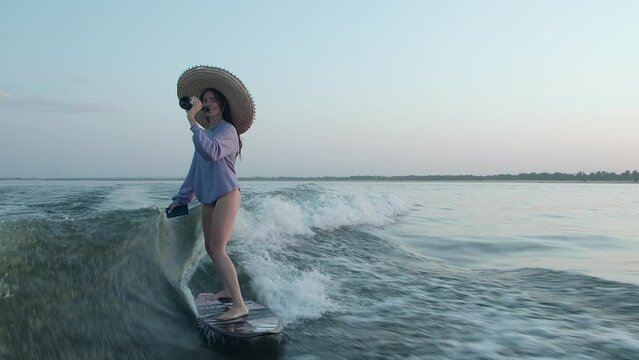 A surfer in a straw hat jumps on a wakeboard with champagne and a phone in her hands. An experienced wakeboarder sprays water drops into the camera.