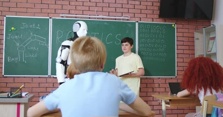 Schoolboy with futuristic white robot standing near blackboard and waving hands to classmates. Young boy controlling humanoid with help of digital tablet. Artificial intelligence. - Powered by Adobe