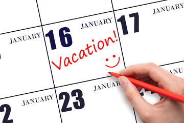 A hand writing a VACATION text and drawing a smiling face on a calendar date 16 January. Vacation planning concept.