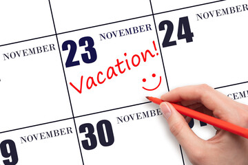 A hand writing a VACATION text and drawing a smiling face on a calendar date 23 November. Vacation planning concept.