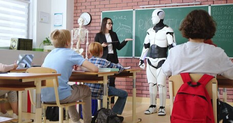 Caucasian female teacher with tablet in hands presenting humanoid robot to attentive pupils at classroom. Interactive lesson at modern school. - Powered by Adobe