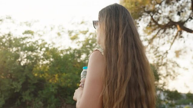 A girl in sunglasses stands against the background of trees and drinks water. On the Sunset. the camera revolves around her