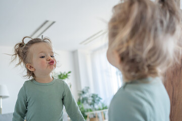Cute funny child girl making funny faces at the mirror and looking at her reflection. Happy...