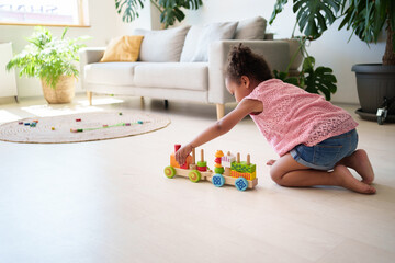 Little black attentive child girl playing wooden eco friendly railroad on the floor. Early learning...
