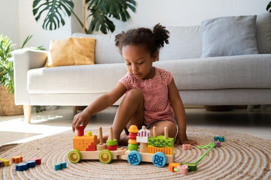 Little black girl engaged in game, playing with wooden building railway station, resting on heating floor at home, enjoying comfort, leisure. African american child playtime and activity concept