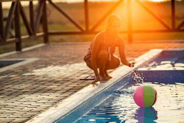 A girl with hair braided in a bun in bright suit plays by the pool with a ball against the...
