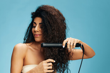 Beautiful enjoyed Latin curly woman using hair straightener, looking down, posing isolated on blue wall background. Hair routine concept, haircare, dry damaged hair ironing, hairdressing. Copy space