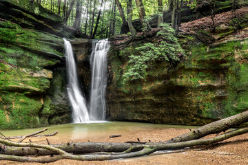 Hidden in a secluded forest, Rock Stalls Falls, a waterfall in the Hocking Hills of Ohio, flows over a sandstone cliff covered in green, with fallen trees in the foreground.