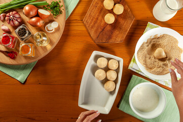 Woman hands preparing brazilian croquette (coxinha de frango) with bread crumbs on a wooden kitchen table - Top view