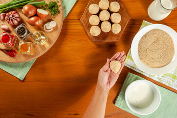 Woman hands preparing brazilian croquette (coxinha de frango) with bread crumbs on a wooden kitchen table - Top view